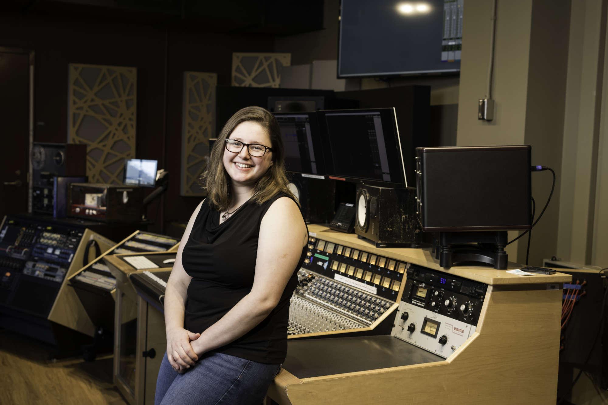 A woman in a black top and jeans leans on the equipment in the control room of a recording studio.