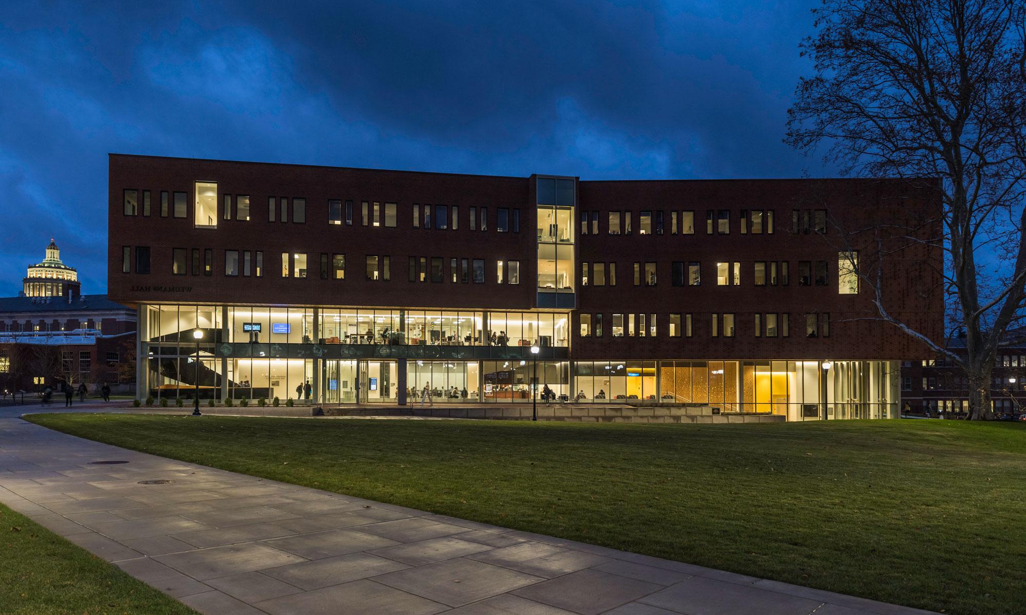 Wegmans Hall, a modern four-story brick building with large glass windows, lit from inside, at dusk. A well-maintained lawn with a walkway is in the foreground, and the University of 罗彻斯特 Library is visible in the background.